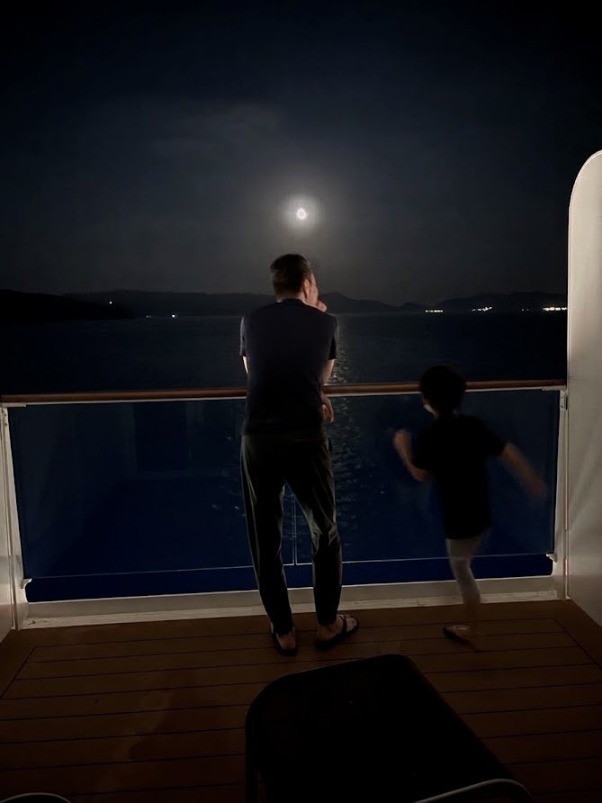 客室のバルコニーから月と夜の海を眺める父と子